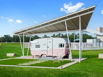 Retro pink-and-white travel trailer parked under a modern metal canopy on a manicured green lawn with stepping stones, white gravel border, two outdoor chairs and a bright blue sky.