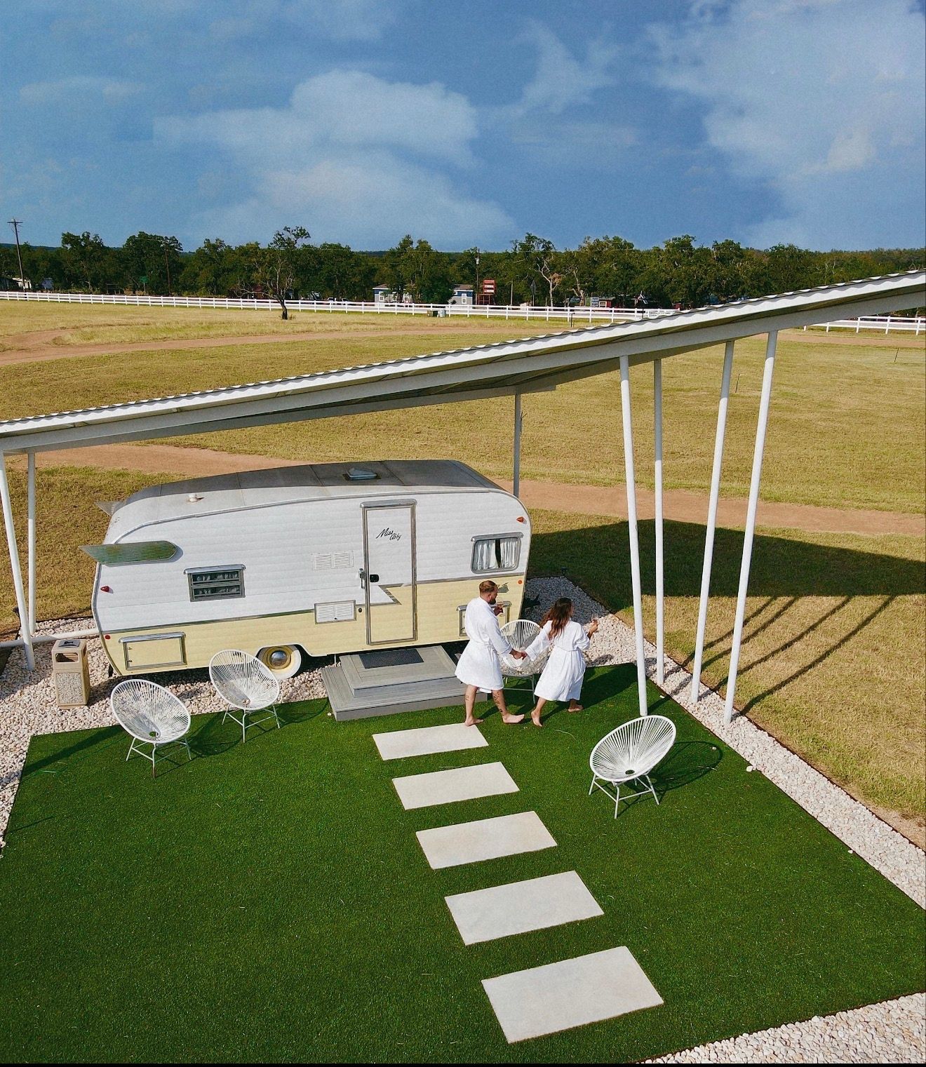 Aerial view of a vintage retro camper glamping setup under a metal canopy on bright green turf with stepping stones and white outdoor chairs, a couple in white robes walking hand-in-hand toward the trailer amid open grassy fields and a white fence.