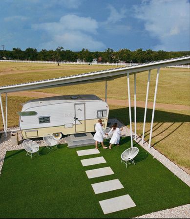 Aerial view of a vintage retro camper glamping setup under a metal canopy on bright green turf with stepping stones and white outdoor chairs, a couple in white robes walking hand-in-hand toward the trailer amid open grassy fields and a white fence.