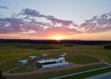 Aerial sunset over rural grassland with a small airstrip, open-sided airplane shelters around a circular dirt track, a modern white building and white fence under pink clouds.