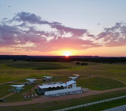 Aerial sunset over rural grassland with a small airstrip, open-sided airplane shelters around a circular dirt track, a modern white building and white fence under pink clouds.