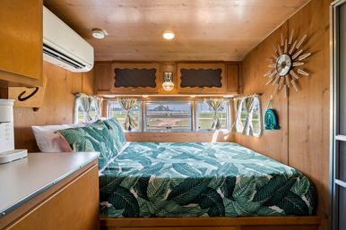 Cozy wood-paneled RV bedroom with queen bed in green tropical leaf bedding, three small windows with tied curtains, overhead storage, wall-mounted air conditioner and retro sunburst clock — compact camper interior
