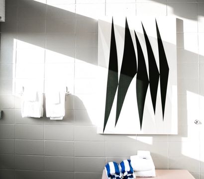 Sunlit modern bathroom with white tiled wall, large black-and-white abstract canvas, diagonal shadows, two white hand towels on hooks and folded blue-striped and white towels on a bench by a frosted glass door.