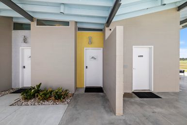 Three white restroom doors beneath a pale-blue covered pavilion with beige and yellow tiled walls, concrete walkways and a small rock garden with green plants.
