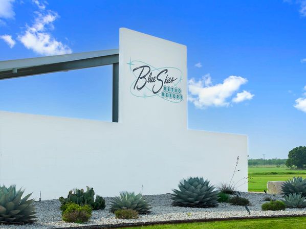 Minimalist white resort entrance sign on a mid-century-style wall beneath a bright blue sky, with agave and cactus in a gravel planting and open green fields beyond.