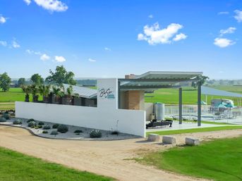 Modern white stucco pool clubhouse with covered patio and fenced swimming pool, palm trees and agave landscaping, dirt driveway and open green fields under a bright blue sky.