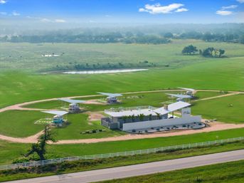 Aerial view of a modern white clubhouse with rooftop pool in a green countryside, circular dirt drive with several colorful canopy-covered tiny cabins, a small pond, white fence and country road under a bright blue sky.