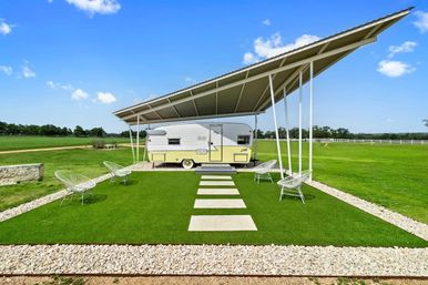 Retro yellow-and-white travel trailer parked under a large angled metal canopy on a manicured green lawn with stepping stones and modern wire chairs, set against a sunny blue sky in a rural field.