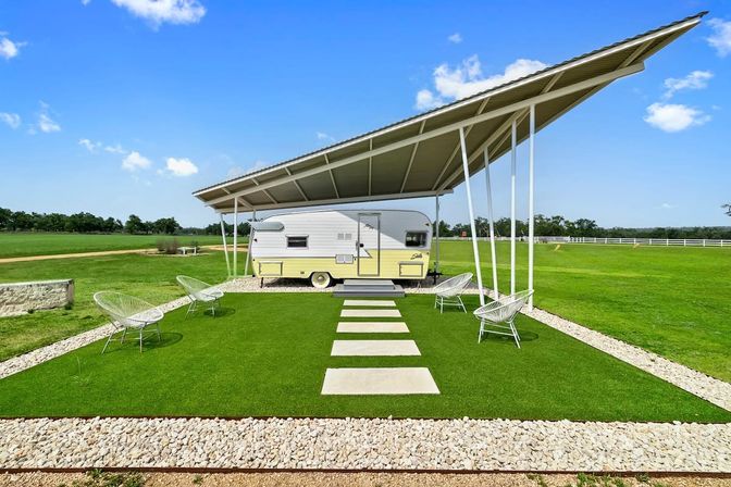 Retro yellow-and-white travel trailer parked under a large angled metal canopy on a manicured green lawn with stepping stones and modern wire chairs, set against a sunny blue sky in a rural field.