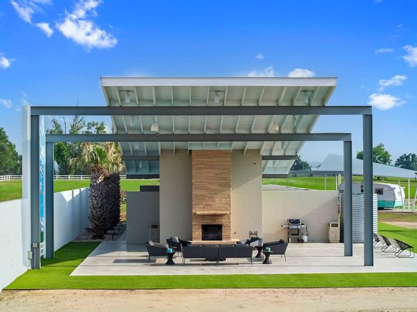Modern covered outdoor pavilion with angled metal roof, central stone fireplace, black lounge seating, grill, palm tree, green lawn and RV under a bright blue sky.
