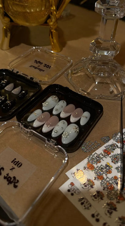DIY Halloween nail art station with trays of embellished press-on nails, a gold skull decoration, crystal candlestick and sheets of Halloween nail decals on a tabletop.