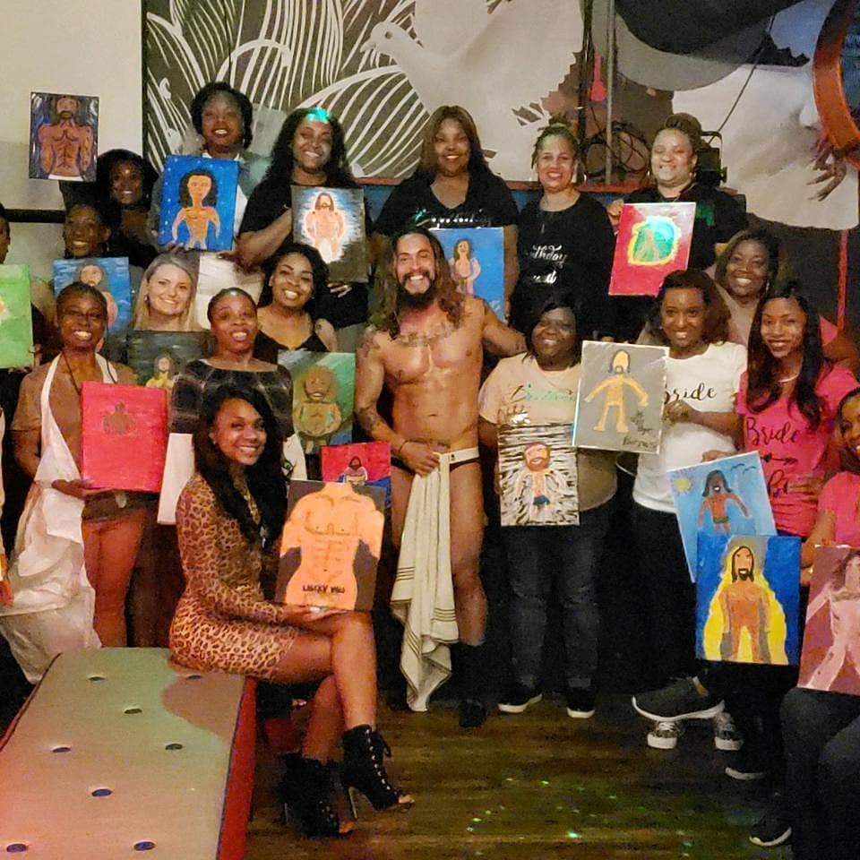 Smiling group of women at a lively paint-and-sip bachelorette party holding canvas paintings of a shirtless male model posing in the center