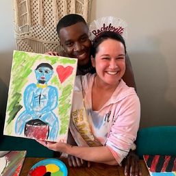 Couple smiling at a bachelorette painting party — woman wearing a bachelorette headband and sash holds a colorful canvas of a blue figure with a red heart, indoor celebration art activity.