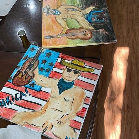 Two colorful folk-art paintings on a wooden table in a home studio: a playful nude cowboy with hat, sunglasses and bandana holding a guitar against an American flag background, and a seated guitarist painting nearby, sunlight on the hardwood floor.