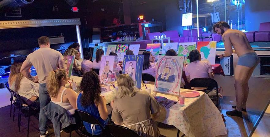 Indoor paint-and-sip style class with people seated at long tables painting canvases of a nearby underwear-clad male model under colorful stage lighting.