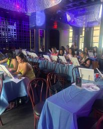 Paint-and-sip class in an urban lounge: people at easels along blue-covered tables, painting beneath crystal chandeliers and purple mood lighting.