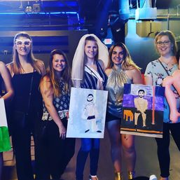 Five women at a bachelorette paint-night in a neon-lit bar or studio, smiling and holding playful paintings of male models; bride-to-be wears a veil and sash.