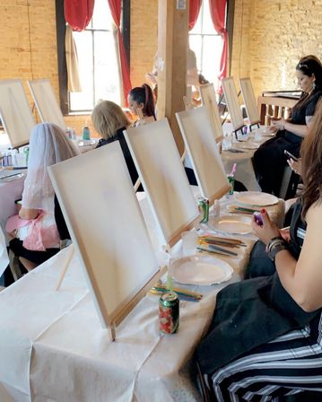 Sunlit brick-walled art studio with rows of easels holding blank canvases, tables laid out with paintbrushes, paper plates and canned drinks, and people seated preparing for a group paint-and-sip session.