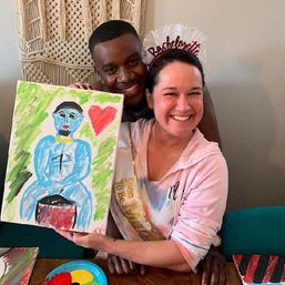 Bride-to-be wearing a bachelorette veil and sash smiles while holding a colorful canvas painting of a blue figure and red heart, friend hugs from behind at an indoor paint-and-sip party with a paint palette on the table.