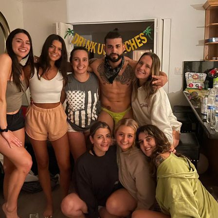 Smiling group of eight friends at a lively house party in a living room, centered on a shirtless man in neon green briefs and a bandana, surrounded by women in casual shorts and hoodies with a decorative party banner and drinks on the counter.