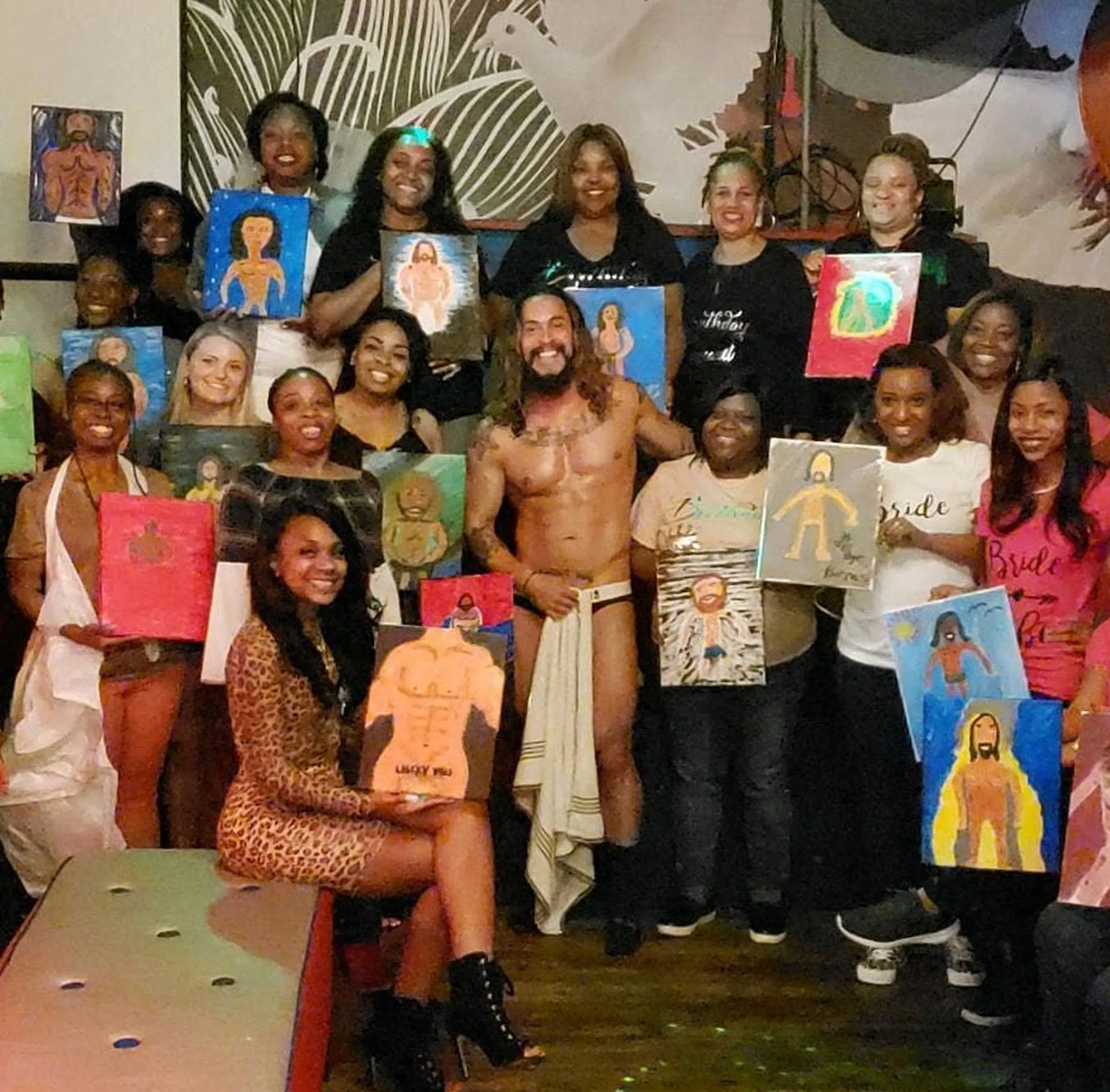 Group of women at an indoor paint-and-sip party posing with colorful figurative canvases and a minimally clad male model in the center, lively bachelorette-style celebration.