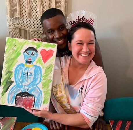Bride-to-be wearing a bachelorette sash and tiara smiles with a friend while holding a colorful DIY canvas painting of a blue figure and red heart at an indoor bachelorette party