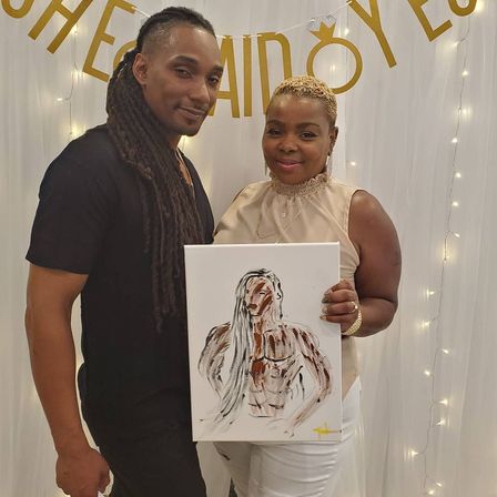 Two adults pose at an indoor celebration holding an abstract painted portrait; white draped backdrop with gold letters and string lights.