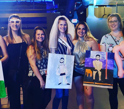 Bachelorette paint-night group: bride-to-be in a veil and friends smiling in a dimly lit studio/bar, holding playful canvas paintings of a male model