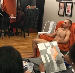 Nude male model seated on an orange-draped chair posing for a life-drawing class while artists in a bright studio with wooden floors paint his portrait on canvases.