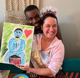 Smiling woman wearing a bachelorette sash and headband with a friend at an indoor paint-night, holding a playful canvas of a blue seated figure and a red heart.