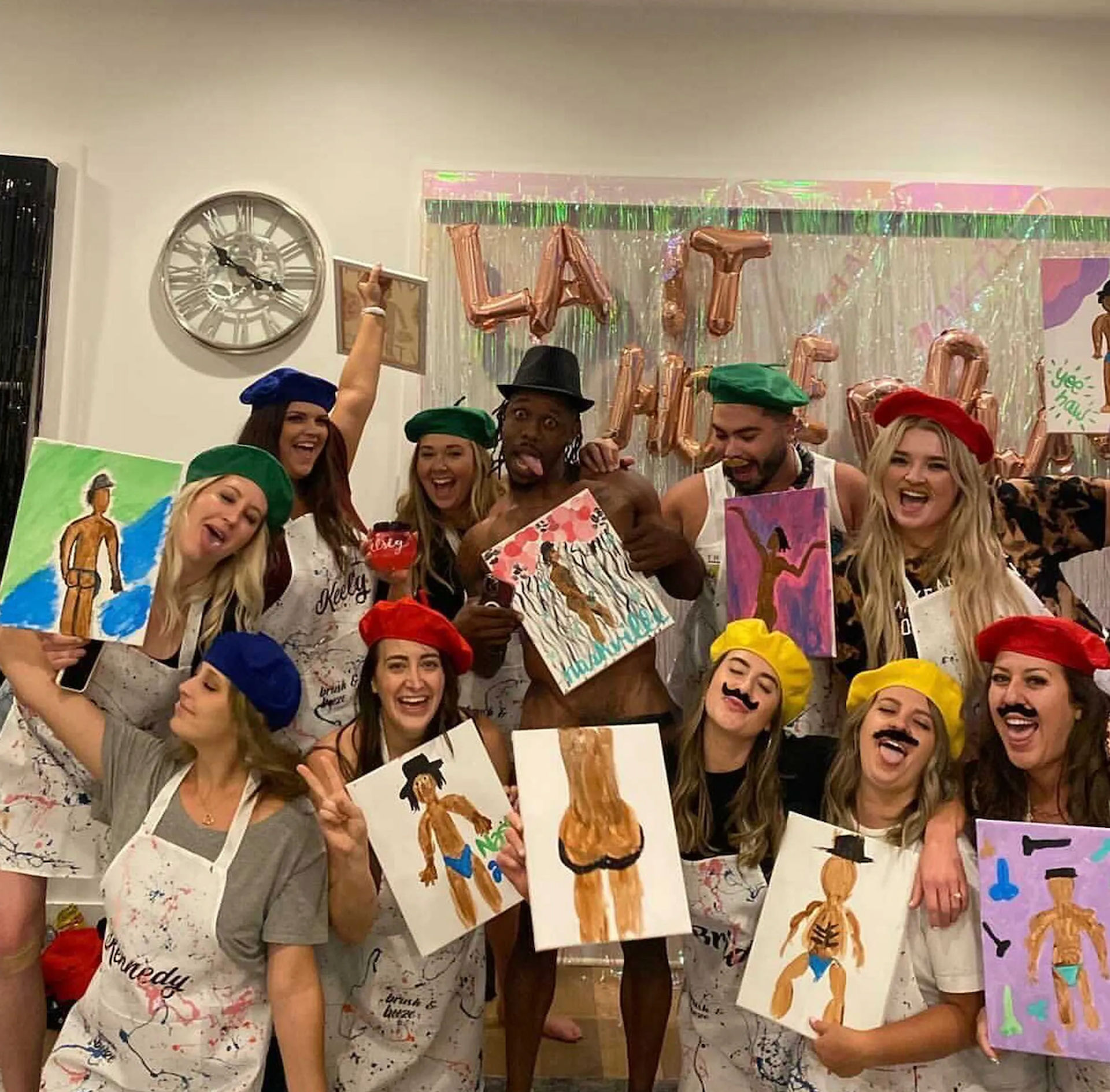 Group of friends at an indoor paint-and-sip party wearing colorful berets and paint-splattered aprons, holding playful figure paintings in front of a metallic balloon backdrop.