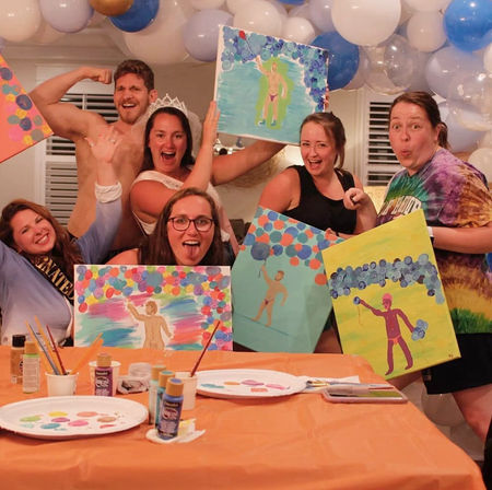 Bachelorette paint night at home: laughing friends and a shirtless man posing under blue-and-white balloon garland, holding colorful DIY canvases with paint bottles and palettes on an orange-covered craft table.