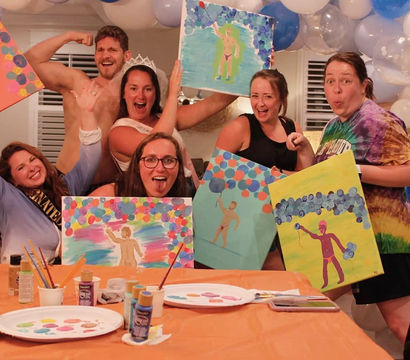 Lively indoor bachelorette paint night with friends posing playfully, holding colorful canvases of a swimsuit-clad figure, paint supplies on an orange-covered table and blue-and-white balloon garland overhead.