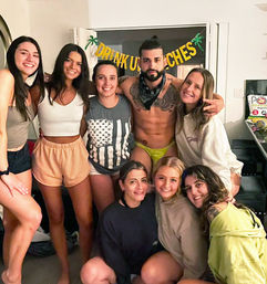 Indoor house party group photo of seven smiling women and a shirtless tattooed man wearing neon swim briefs and a bandana, posing in a living room under a festive party banner