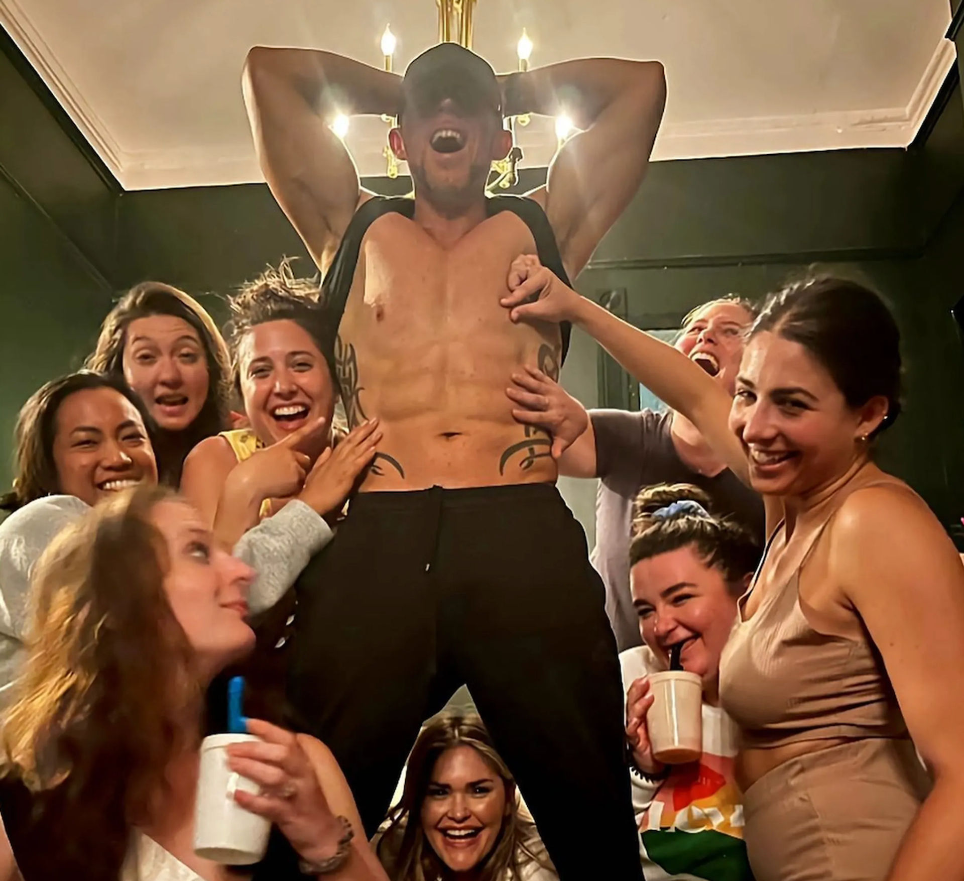 Group of women at a lively indoor party crowd a shirtless man standing with arms raised under a chandelier, laughing and posing in a living room.