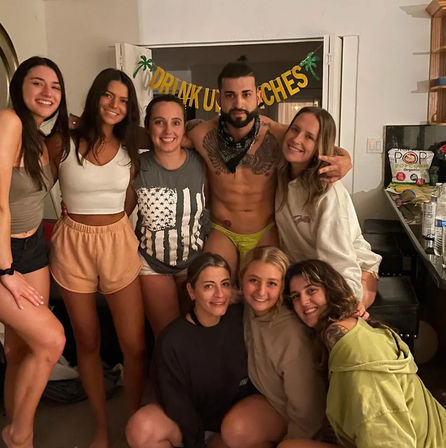 Smiling crew of eight friends posing at a casual indoor house party in a living room/kitchen; shirtless man in a neon swim brief and bandana at center, friends in shorts and sweatshirts, festive tropical banner and snacks on the counter.