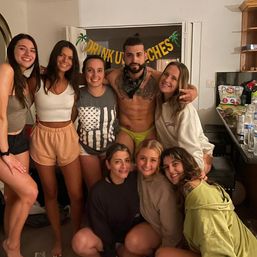 Eight friends posing at a casual indoor house party in a living room — shirtless man in neon briefs and women in loungewear, tropical party banner overhead and drinks and snacks on the counter.