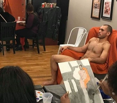 Male nude model reclining on an orange-draped chair in a life-drawing class while artists sketch and paint in a studio