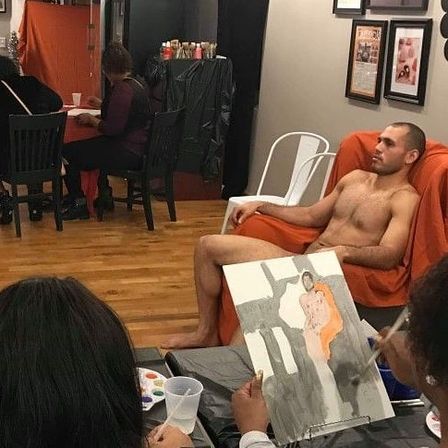 Shirtless model reclining on an orange chair in a hardwood-floor art studio while artists at easels paint a life-drawing portrait with gray and orange tones.