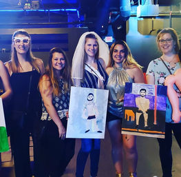 Smiling group of women at a bachelorette paint-and-sip night in a dimly lit bar; bride in a veil and sash holds a painted canvas while friends display colorful portrait paintings.