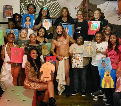 Bachelorette paint-and-sip group indoors, smiling women holding canvases of a shirtless male model who stands center with a towel, festive and playful vibe