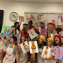 Indoor paint-and-sip party with a lively group wearing colorful berets and paint-splattered aprons, holding humorous canvases of male figures and posing with a shirtless model in front of festive balloons and a wall clock.