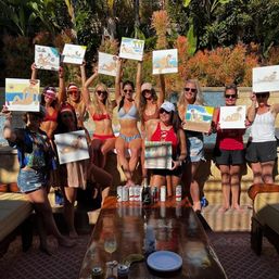 Sunny backyard poolside sip-and-paint: group of women in swimwear holding up beach-themed canvases with drinks and snacks on a table