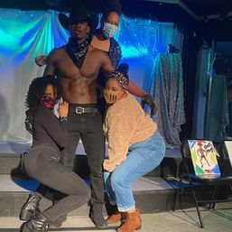 Shirtless male performer in a cowboy hat and bandana poses on a small theater stage under blue-green lights with three masked women crouching around him in a playful group photo.