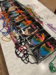 Mardi Gras-inspired rainbow sugar-coated pastries in clear plastic boxes lined up on a marble countertop, surrounded by colorful bead necklaces