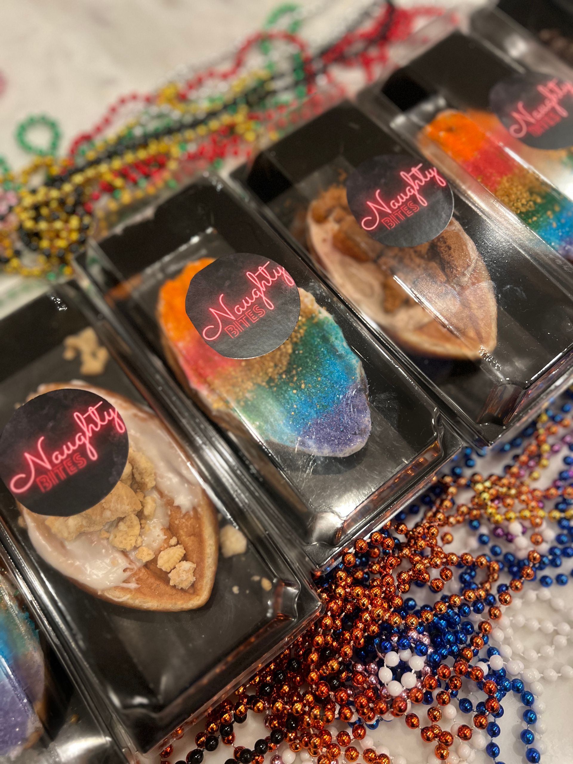 Clear plastic boxes of mini glazed pastries and rainbow-sprinkled treats arranged as party favors on a table scattered with orange, blue, gold and green Mardi Gras beads, each box topped with a black sticker and pink script.