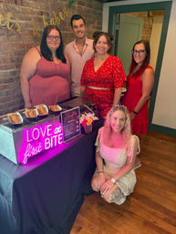 Smiling group of five at an indoor dessert station with exposed brick wall and gold banner, neon pink "LOVE at first BITE" sign over waffle cones; bride-to-be wearing a tiara kneels in front.
