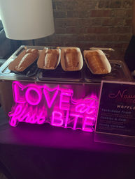 Four waffle-pastry sandwich treats in paper trays on a metal display against a brick wall, lit by a bright pink neon sign reading "LOVE at first BITE".