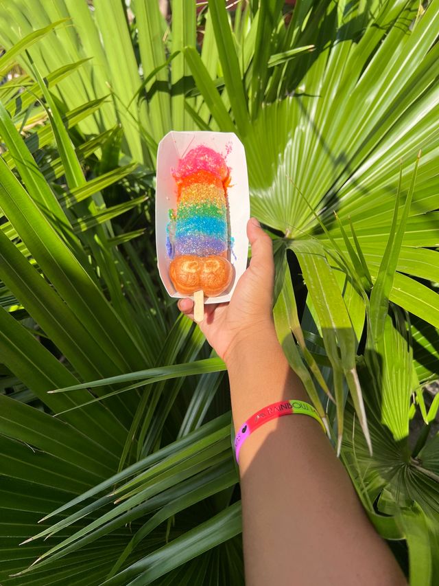 Hand holding a rainbow-sprinkled ice cream bar on a wooden stick in a white tray over lush tropical palm leaves, bright summer dessert photo