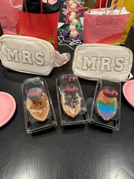 Bachelorette party table with two white cosmetic pouches embroidered MRS, colorful gift bags and beads, and three assorted decorated pastries in clear boxes on a black surface.